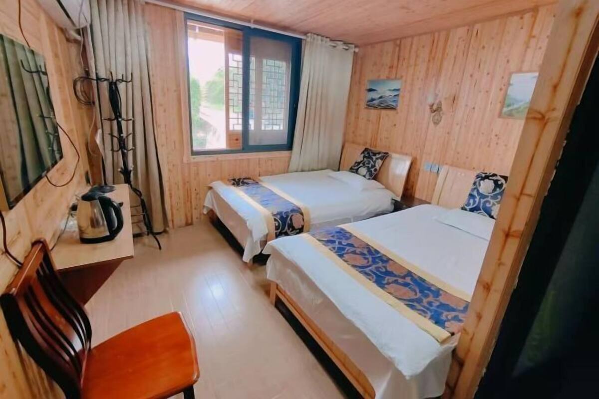 Ein Zimmer mit zwei Betten, einem Holztisch, einem Stuhl und einem Fenster mit Vorhängen.