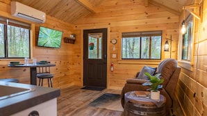 Een gezellig interieur van een chalet, met houten details, een eethoek met tafel en stoelen, een bank met een plant, en een aan de muur bevestigde televisie.