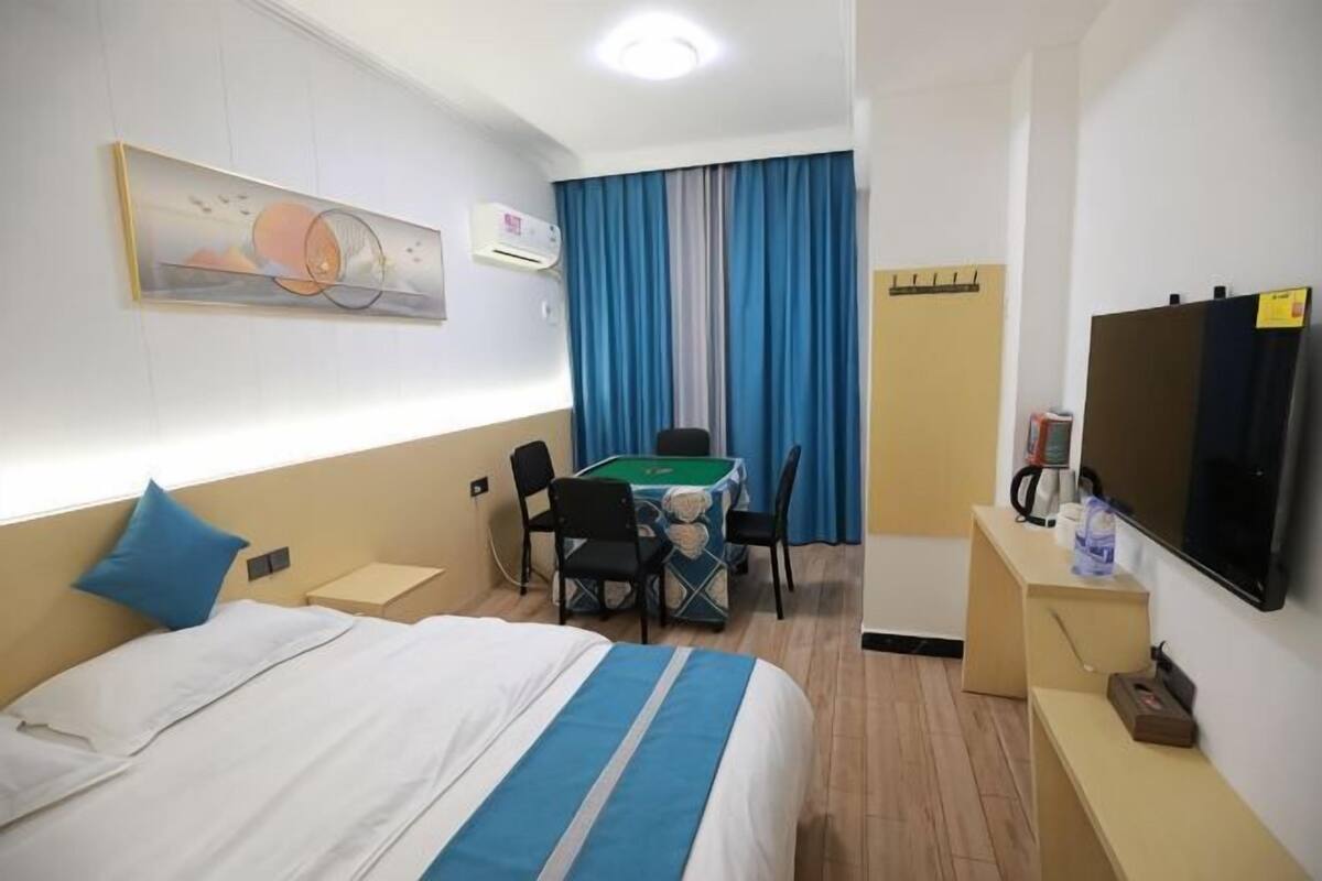 A hotel room with a bed, a desk with a green table, a TV, and a window with blue curtains.