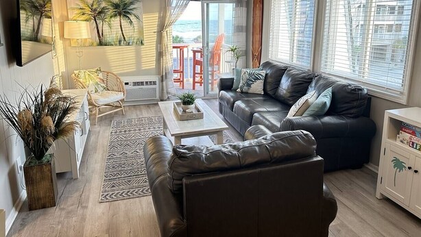Een woonkamer met een bank, salontafel en stoelen. Er is een groot raam met zonwering, een televisie en een schilderij van palmbomen.