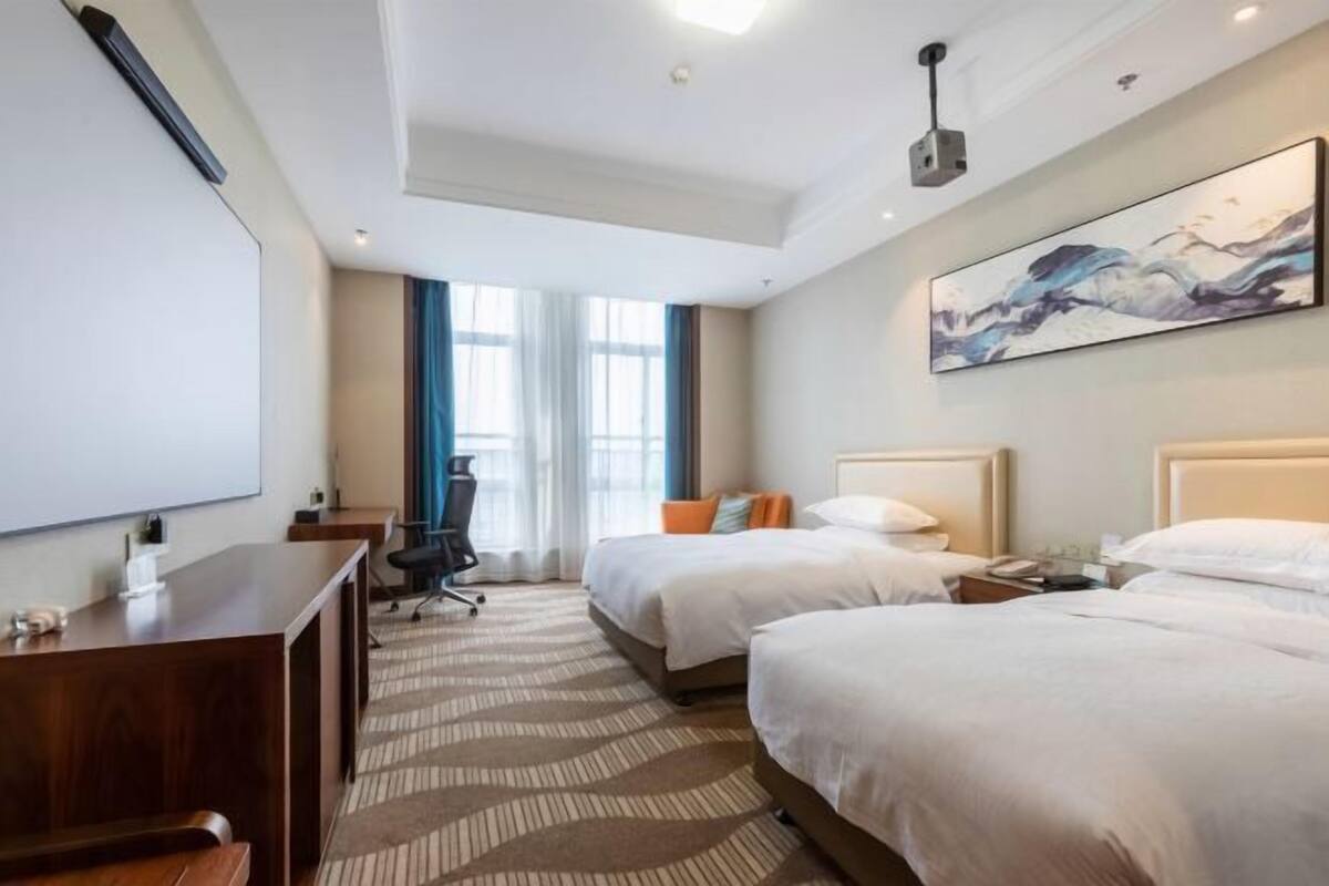 Une chambre d’hôtel avec deux lits, un bureau, une chaise, une grande fenêtre avec des rideaux et un tableau au mur.