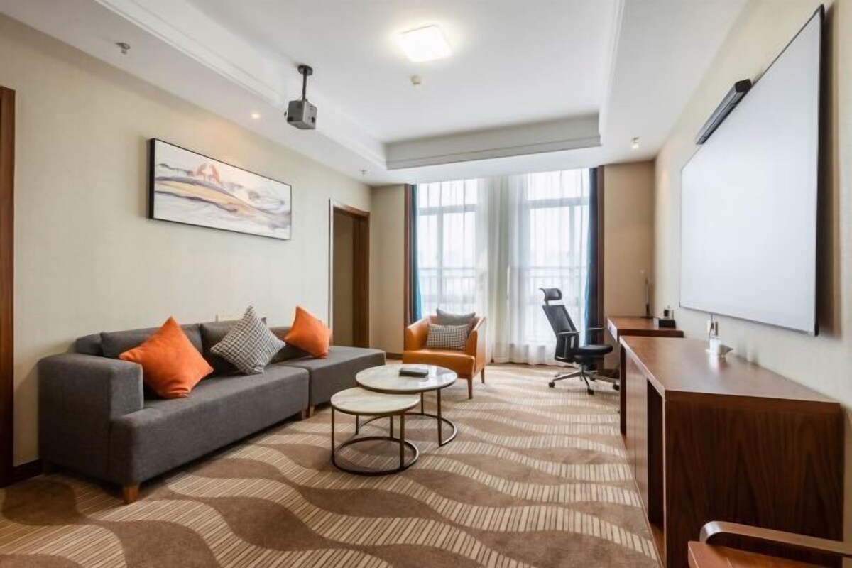 A modern hotel room with a sofa, armchair, coffee table, and a large screen.