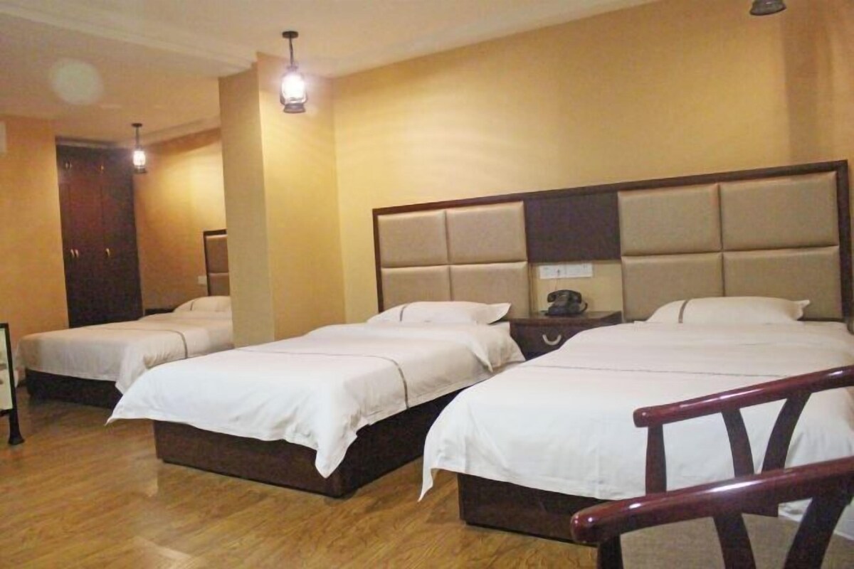 A hotel room with three single beds, wooden flooring, a large padded headboard, and a small bedside table with a lamp.