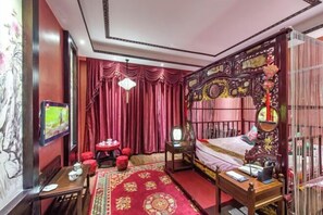 Una habitación tradicional con cama, escritorio y silla. Hay una cortina roja y un cuadro en la pared.