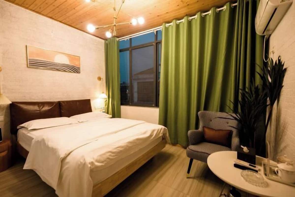 Un dormitorio con cama, una silla, una mesita, una lámpara, una ventana con cortinas y un aire acondicionado de pared.