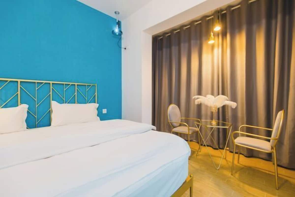 Habitación de hotel con una cama grande, una pared de acento azul, una cortina y una pequeña mesa con sillas.