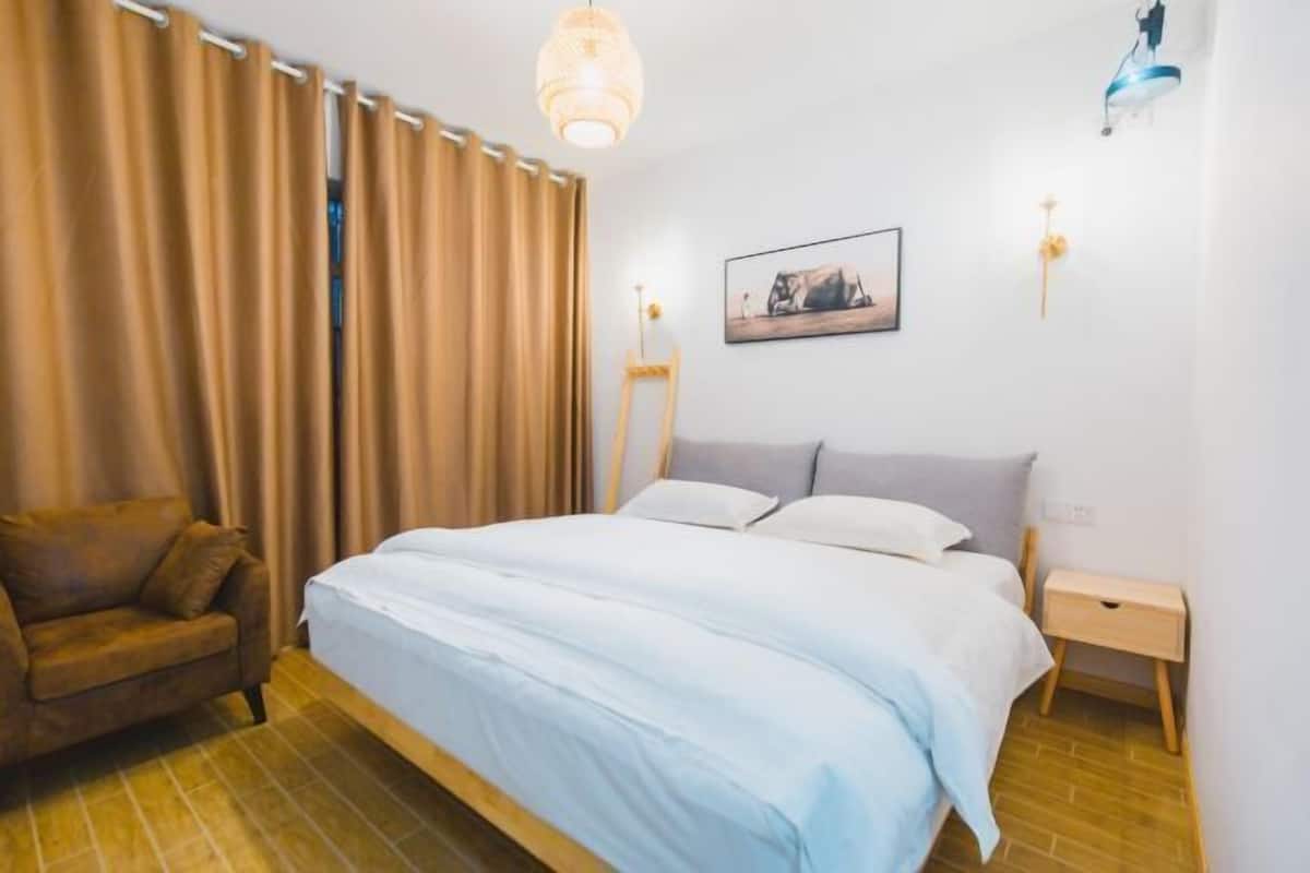 Un dormitorio con cama, una silla, una mesita de noche, un cuadro en la pared y una lámpara colgante.