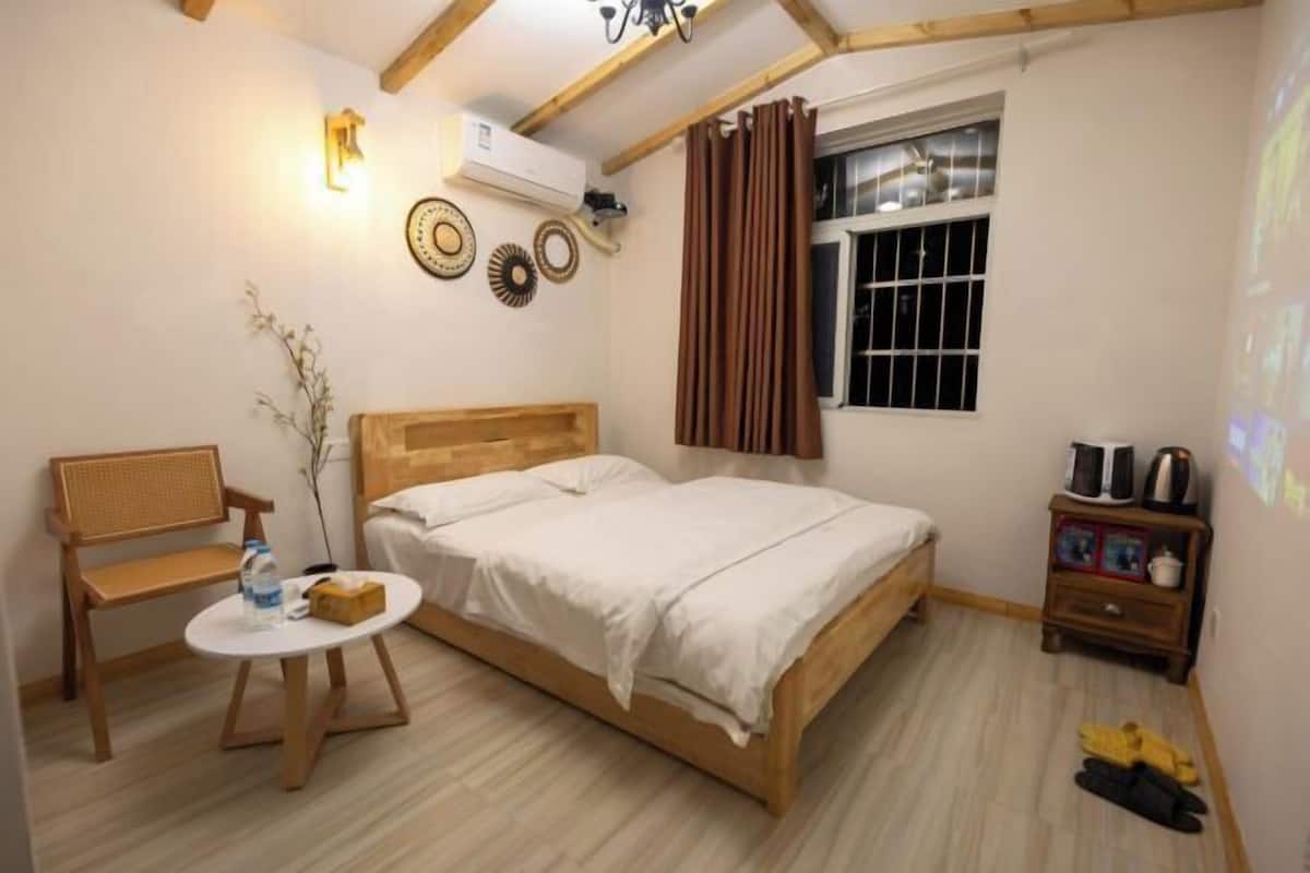 Un dormitorio con cama, una mesita con una botella, un banco, una mesita de noche con tetera y una ventana con cortinas.