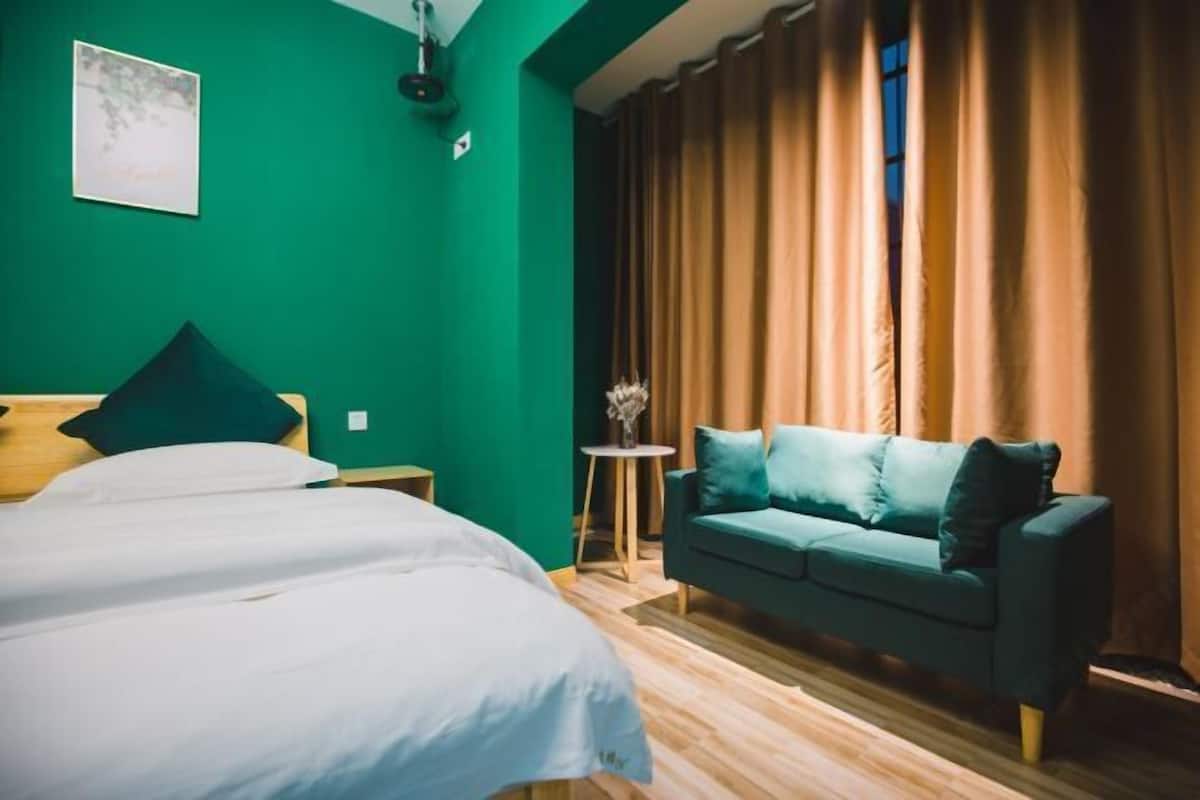 Una habitación con una cama, un muro verde, un sofá verde, una mesita con un jarrón y una ventana con cortinas.