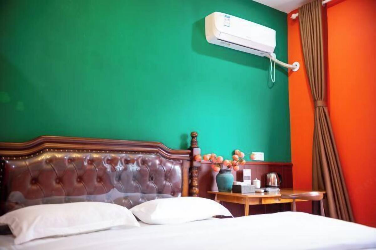 Una habitación de hotel con cama, cabecera de madera, mesita de noche con un florero y un equipo de aire acondicionado en la pared.