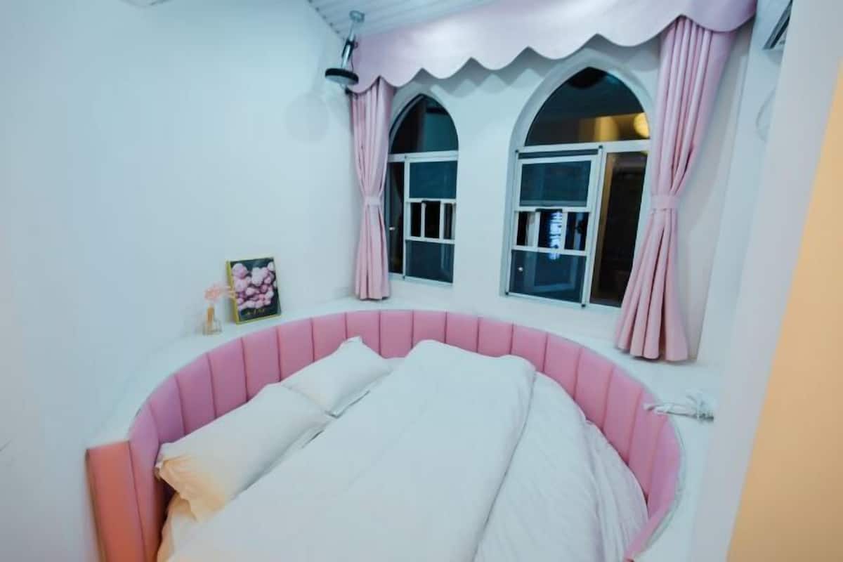 Un dormitorio con una cama tapizada en rosa, dos ventanas con cortinas rosadas y una mesita de noche con un cuadro enmarcado.