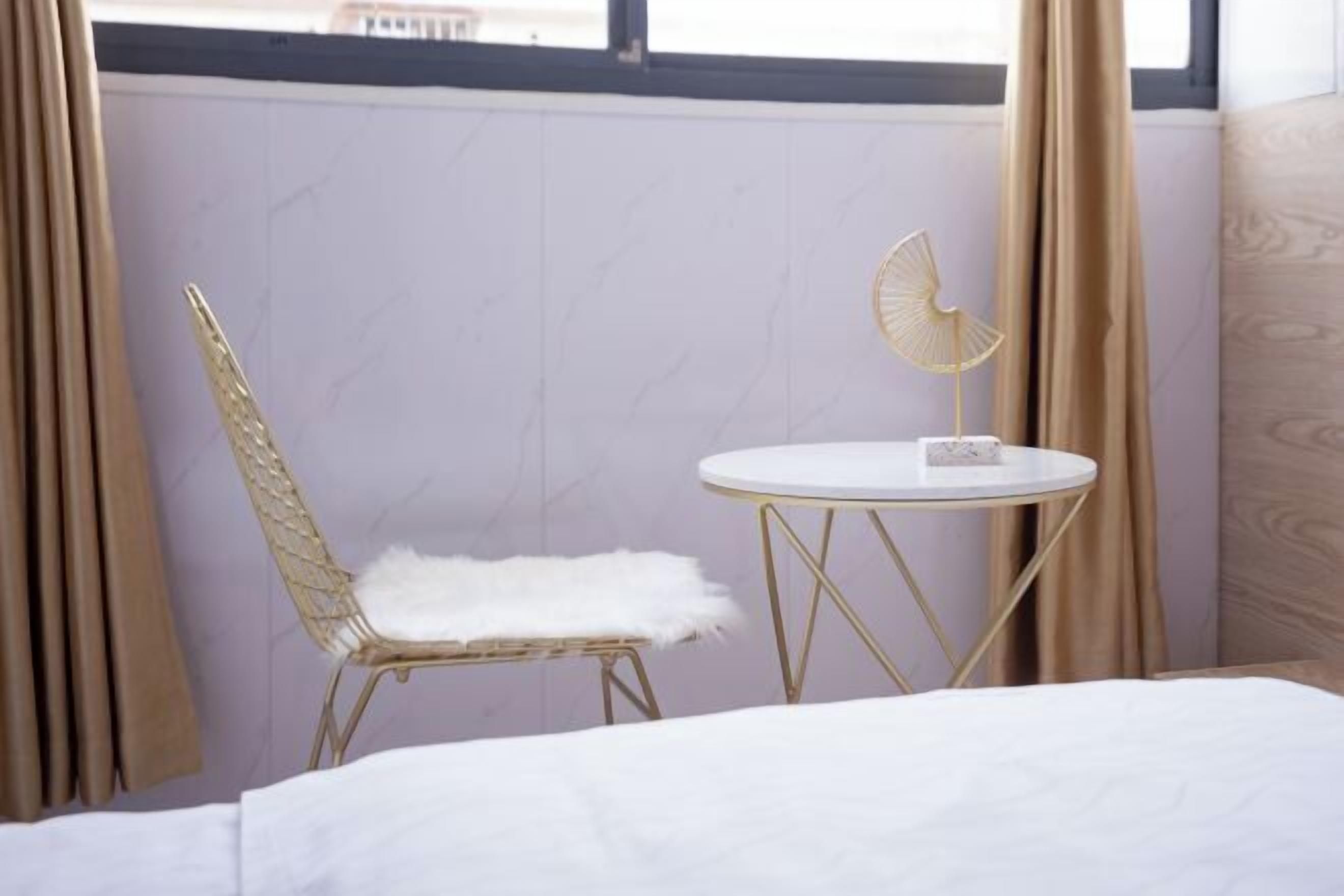 Una habitación con una cama, una mesa redonda con cubierta blanca, una silla dorada con cojín blanco y un abanico dorado sobre la mesa.