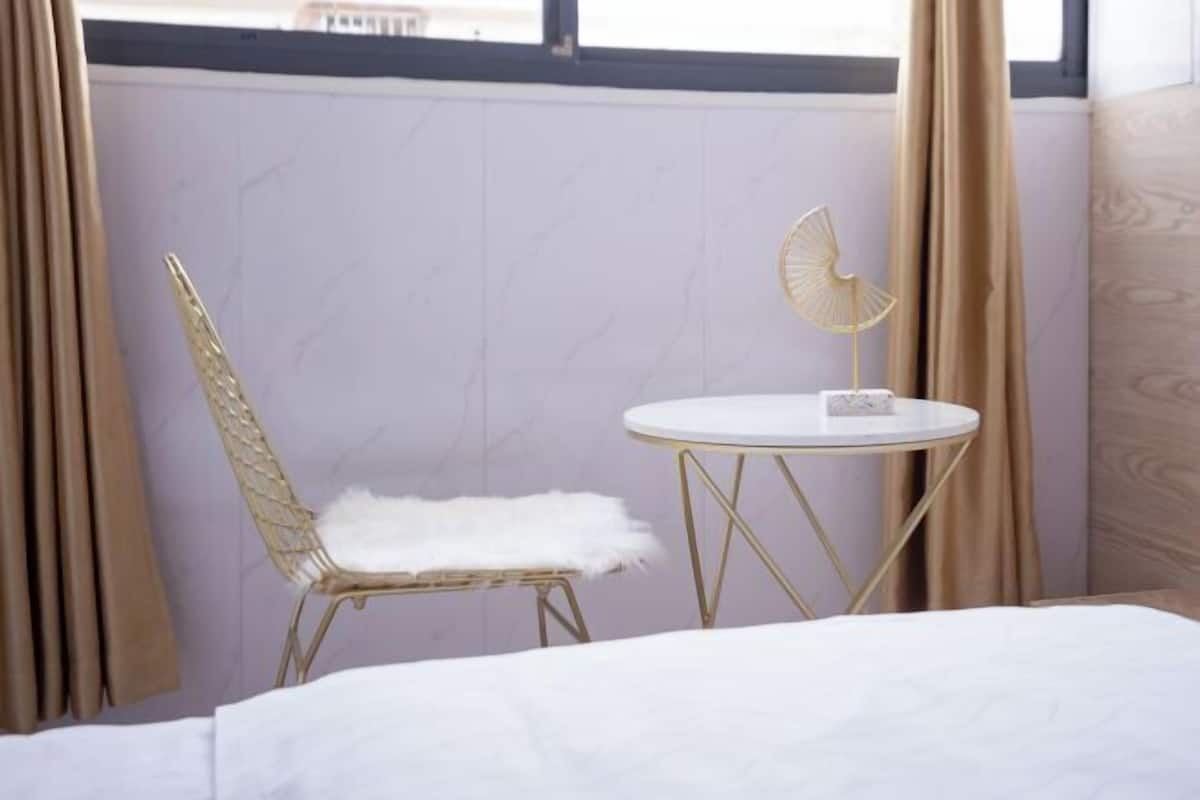 Una habitación con una cama, una mesa redonda con cubierta blanca, una silla dorada con cojín blanco y un abanico dorado sobre la mesa.