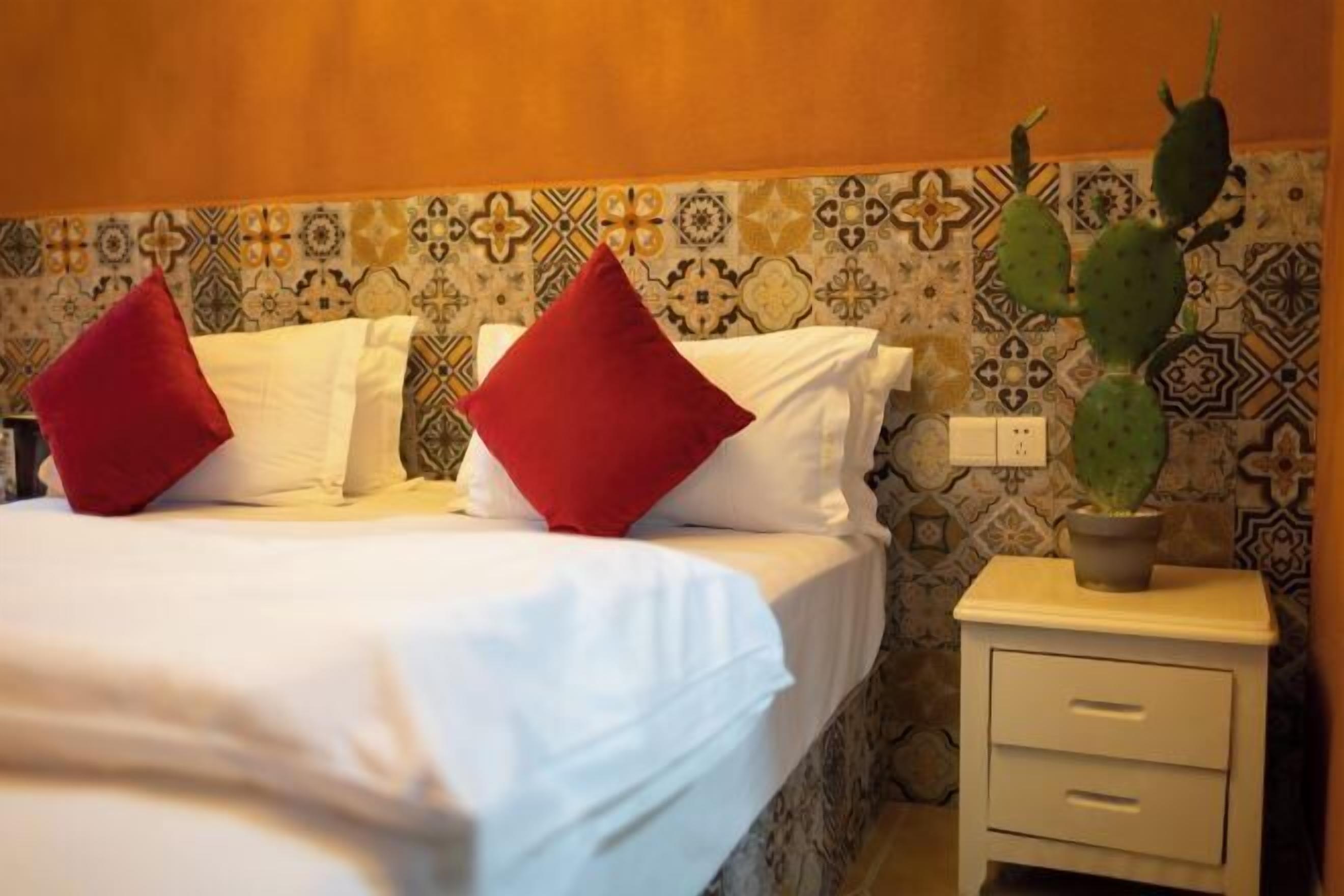 Habitación de hotel con cama, dos almohadas rojas, mesita de noche con planta y papel tapiz estampado.