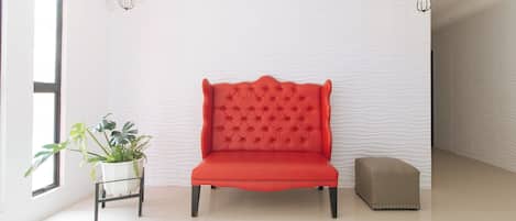 Una butaca roja con tapizado, en una habitación con paredes blancas y texturizadas, una planta en maceta y un taburete pequeño.