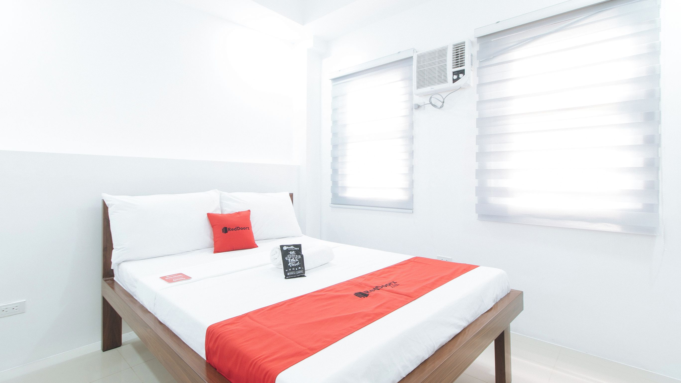 Habitación de hotel con una cama, paredes blancas, una ventana con persianas y una almohada roja.