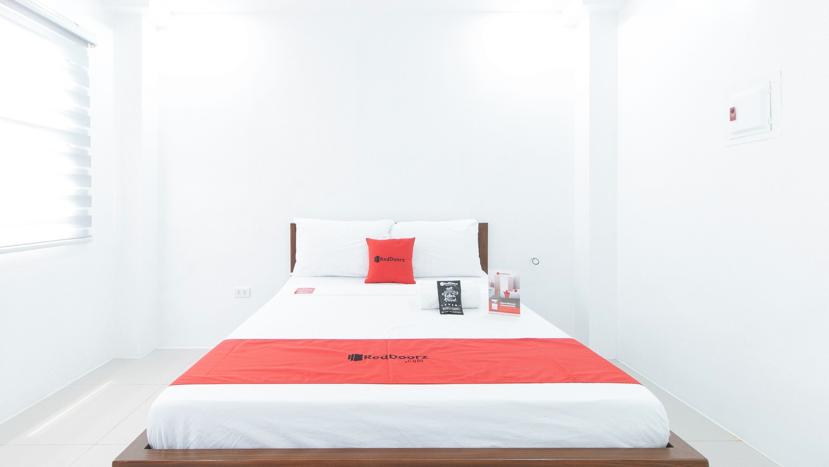 Una cama bien tendida con una colcha blanca y roja, un cojín rojo y un pequeño folleto a un lado.