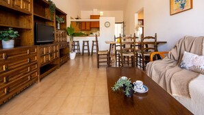Ein Wohnzimmer mit einer Holzeinrichtungsanlage, einem Fernseher und einem Couchtisch mit einer Pflanze. Es gibt einen Essbereich mit einem Holztisch und Stühlen, und im Hintergrund eine Küche.
