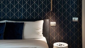 A modern hotel room with a geometric patterned wall, a bed with white and dark blue bedding, a bedside table with a lamp, and a wall-mounted light fixture. - Rubini 7 Suite Gold - MyHomeinComo (Como)