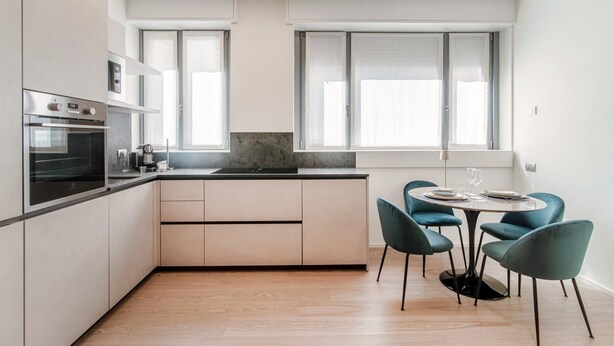 A modern kitchen with built-in appliances, a dining area with teal chairs, and large windows. - Rubini 7 Suite Gold - MyHomeinComo (Como)