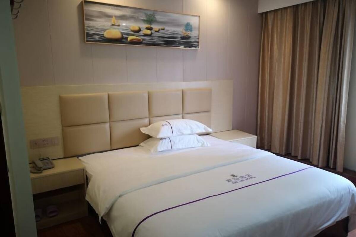 Una habitación de hotel con una cama grande, cabecero, mesita de noche, un cuadro en la pared y una ventana con cortinas.