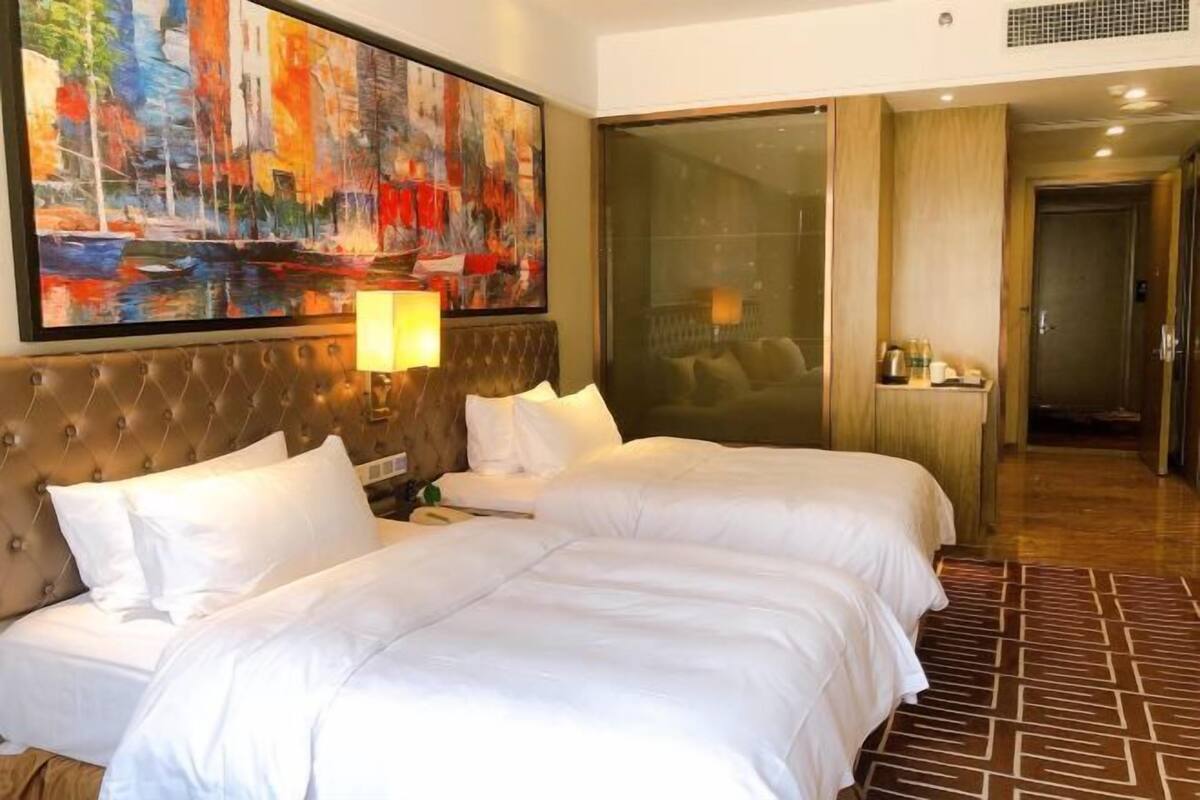 Une chambre d’hôtel avec deux lits, un grand tableau, une porte vitrée donnant sur un balcon et une salle de bain visible.