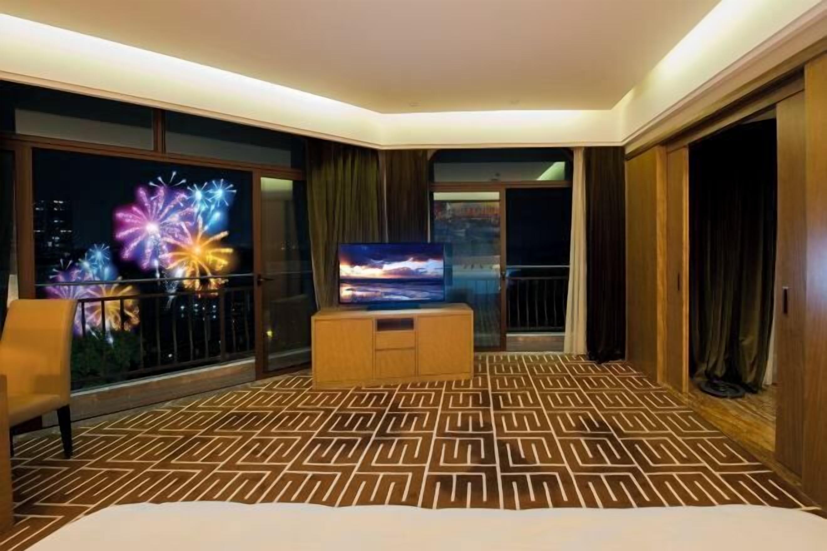 A hotel room with a large bed, a TV, a balcony with a view of fireworks, and a patterned carpet.