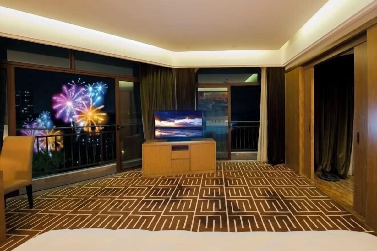 A hotel room with a large bed, a TV, a balcony with a view of fireworks, and a patterned carpet.