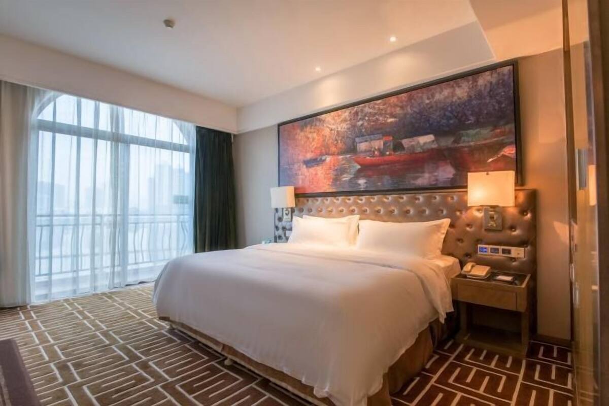 Une chambre d’hôtel avec un grand lit, une tête de lit à motifs capitonnés, une table de chevet avec une lampe et une grande fenêtre avec des rideaux.