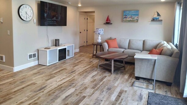 Een moderne woonkamer met een neutraal kleurenpalet, een flatscreen televisie aan de muur, een houten salontafel, een grijze bank met oranje kussens, en een kleine decoratie van een zeilboot aan de wand.