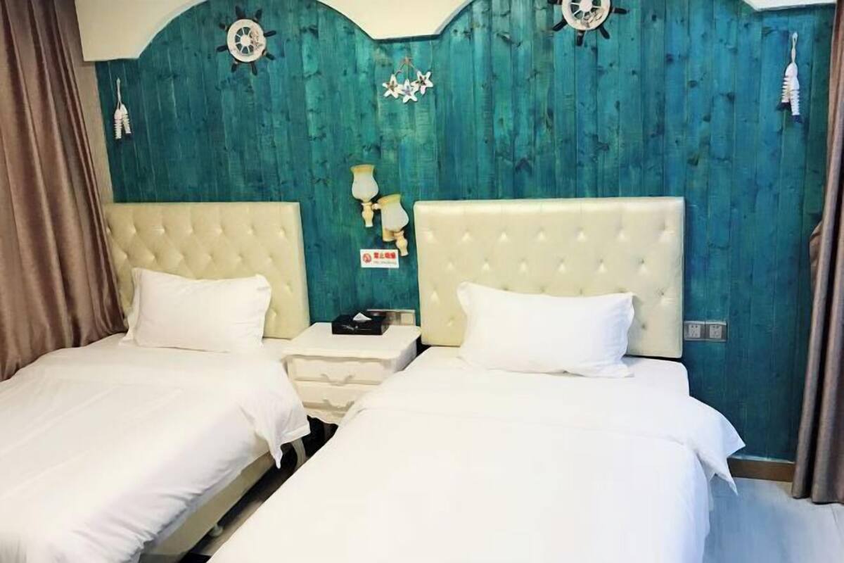 A room with two beds, each with a white headboard and white bedding. There are wall-mounted lamps and a clock on the teal-colored wall.