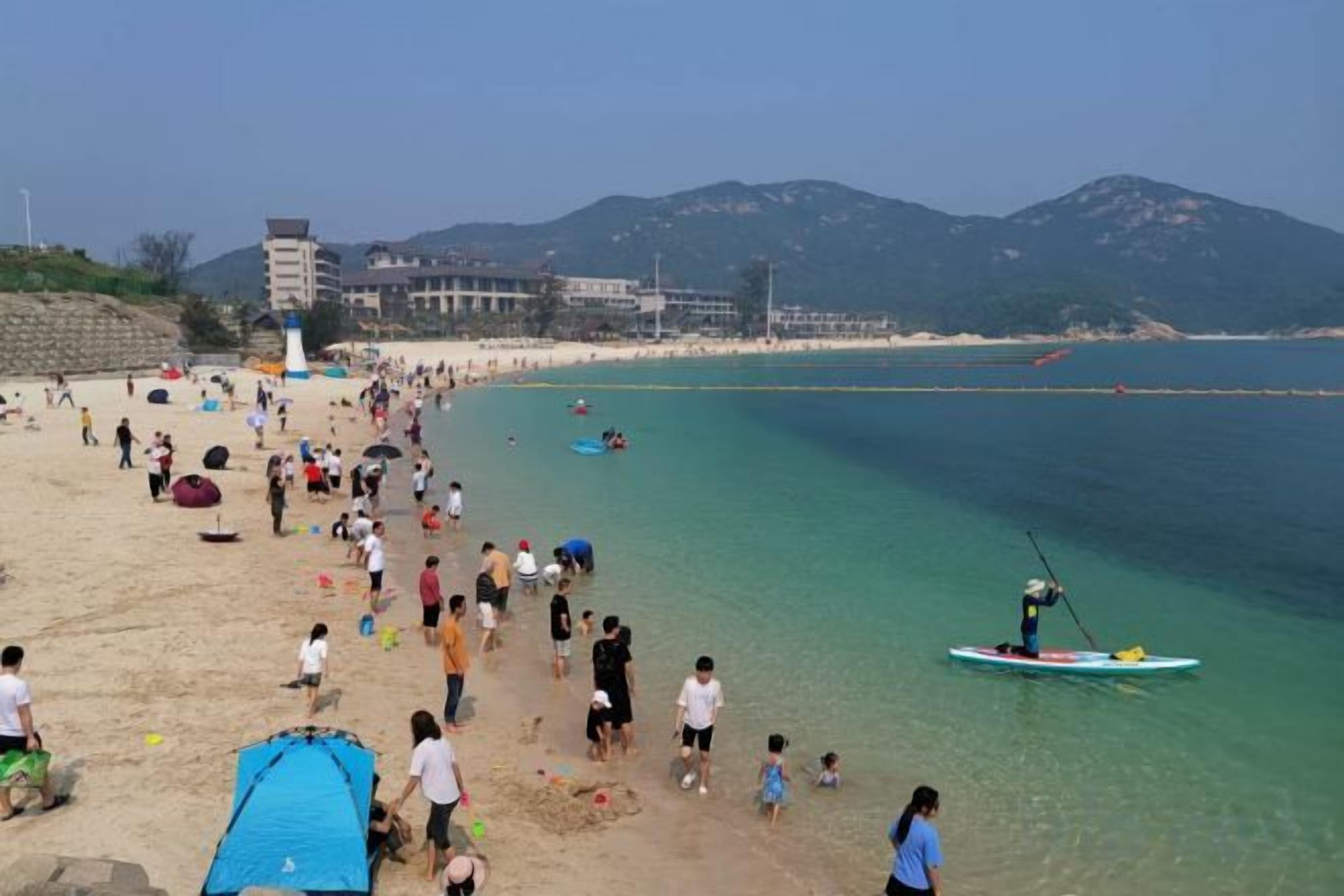 A beach with people, a lighthouse, and mountains in the background.