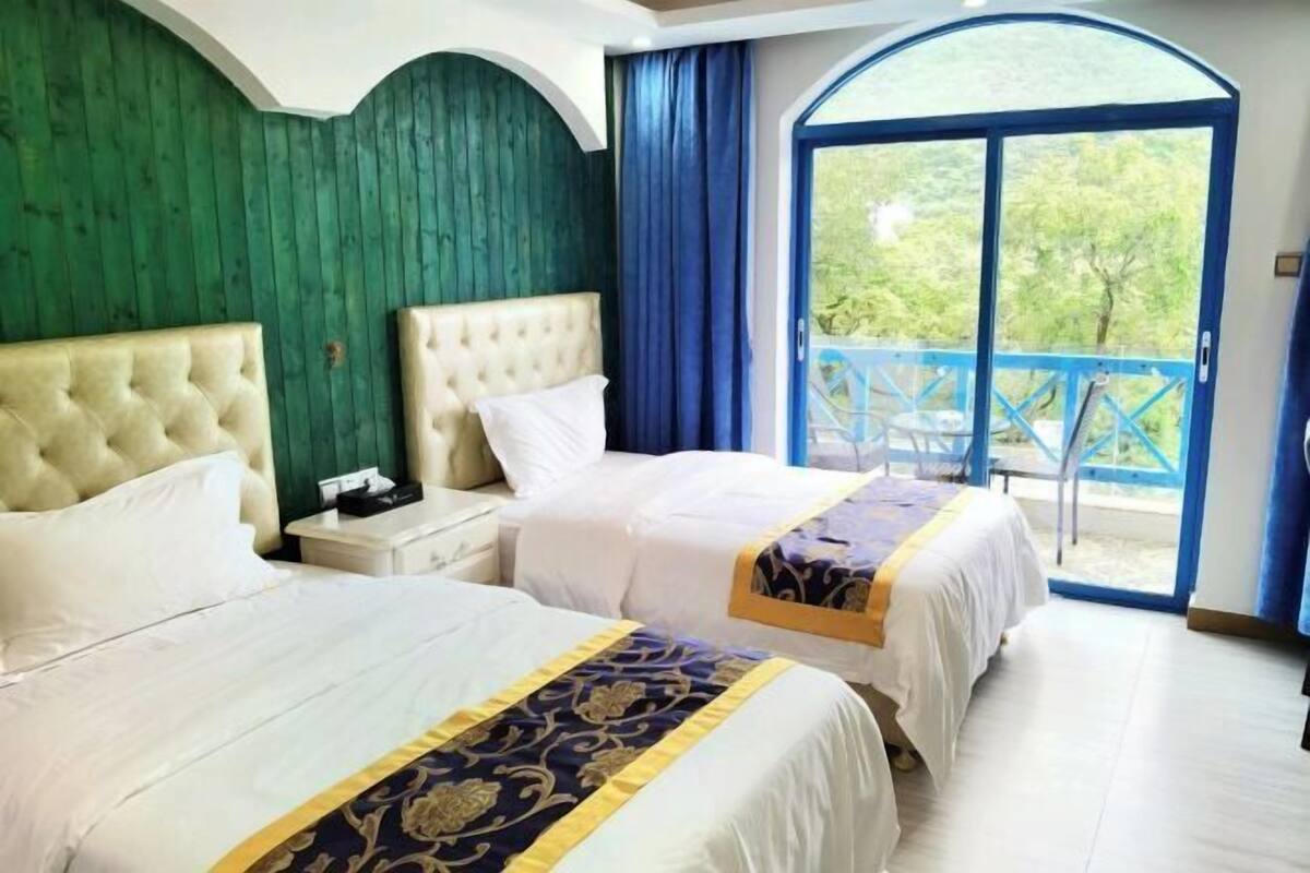 A hotel room with two beds, a large window with a view of greenery, and a balcony with blue railings.