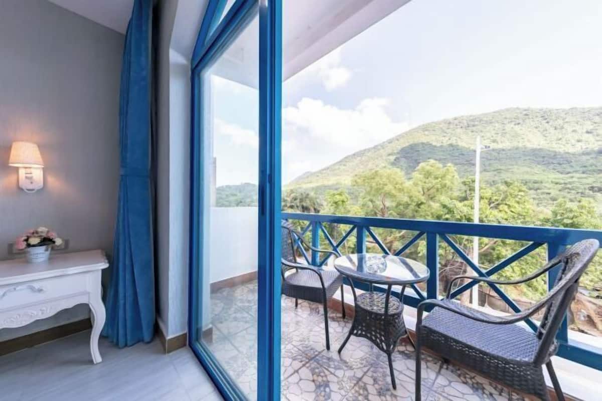 A balcony with a table and chairs, a white console table with a lamp, and a view of a hilly landscape.