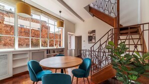 A dining area with a round table and blue chairs, a staircase with a metal railing, and a view of a brick building through large windows. - Apartment with Terrace on the Vatican Passetto (Roma)
