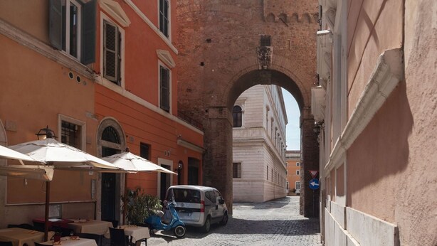 A narrow street with a brick archway, outdoor seating with umbrellas, and a parked car. - Apartment with Terrace on the Vatican Passetto (Roma)