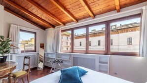 A bedroom with a wooden ceiling, a large bed with blue pillows, a window with a view of buildings, a radiator, and a small table with a chair. - Apartment with Terrace on the Vatican Passetto (Roma)