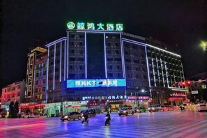 A multi-story hotel with a prominent sign at the top, multiple ground-level shops, and a parking lot with cars.