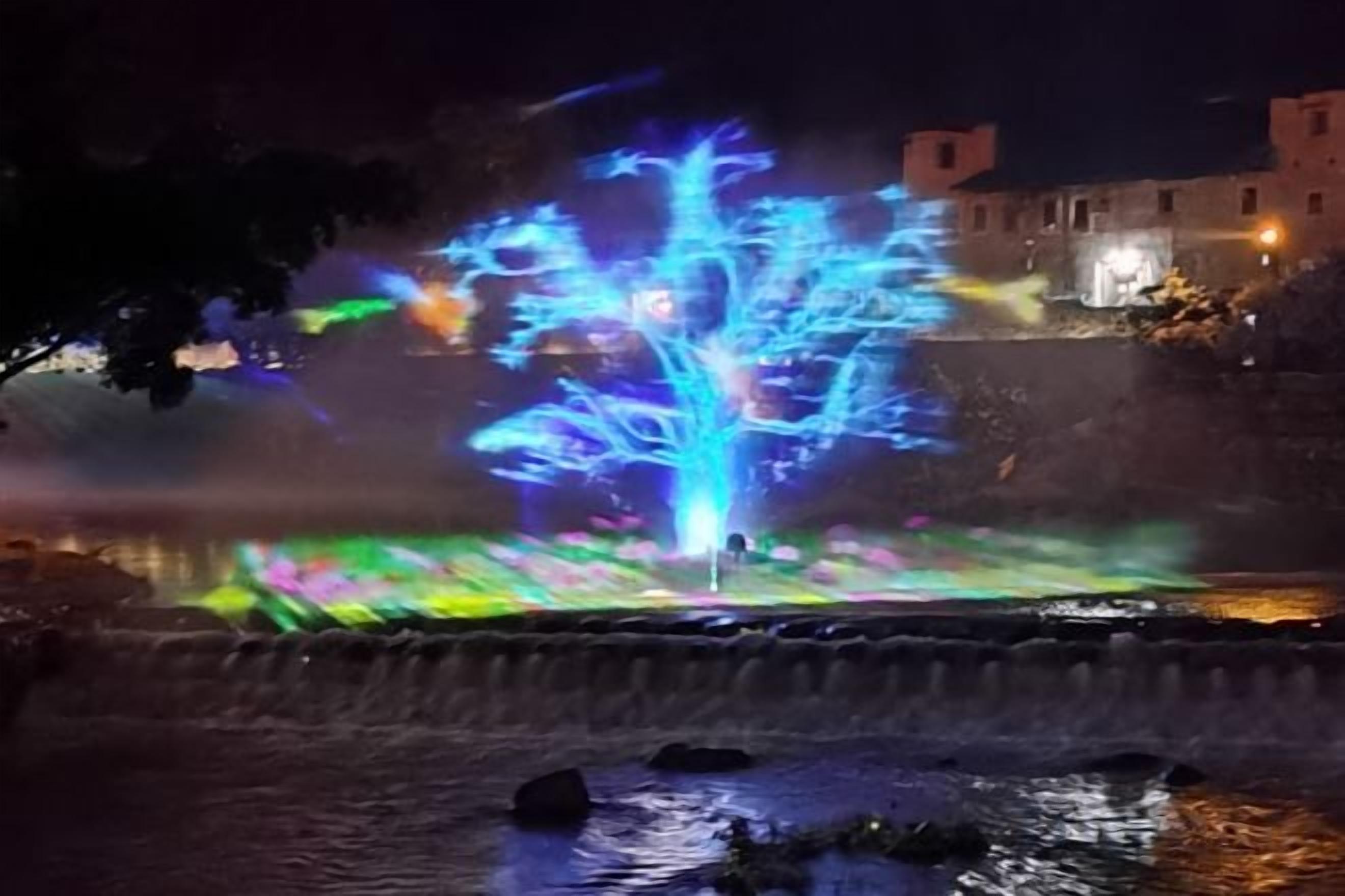 Une vue nocturne d'une installation aquatique illuminée par des jeux de lumière, avec un pont et des bâtiments en arrière-plan.