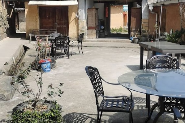 Un patio exterior con una mesa y sillas redondas, un pequeño jardín con plantas y la entrada de un edificio al fondo.