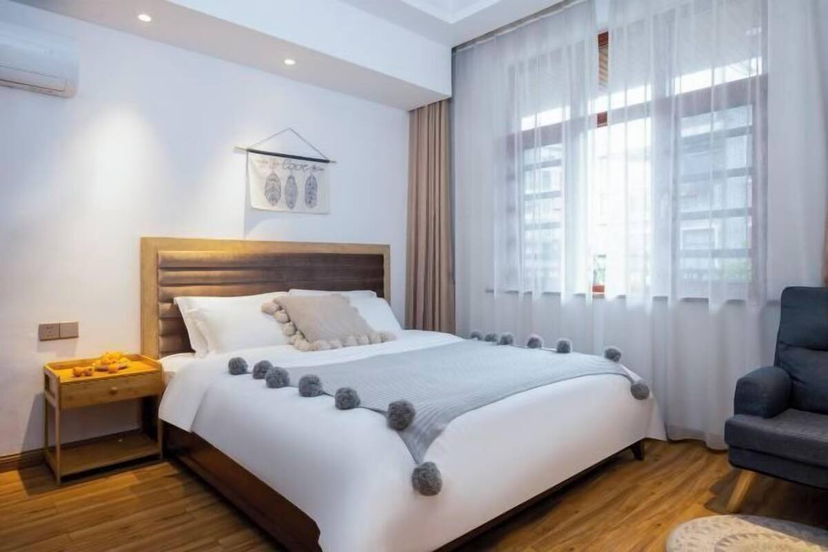 Ein Schlafzimmer mit einem hölzernen Kopfteil, einem Bett mit weißer Bettwäsche und grauen Pompons, einem Nachttisch mit Obsttablett und einem Sessel.