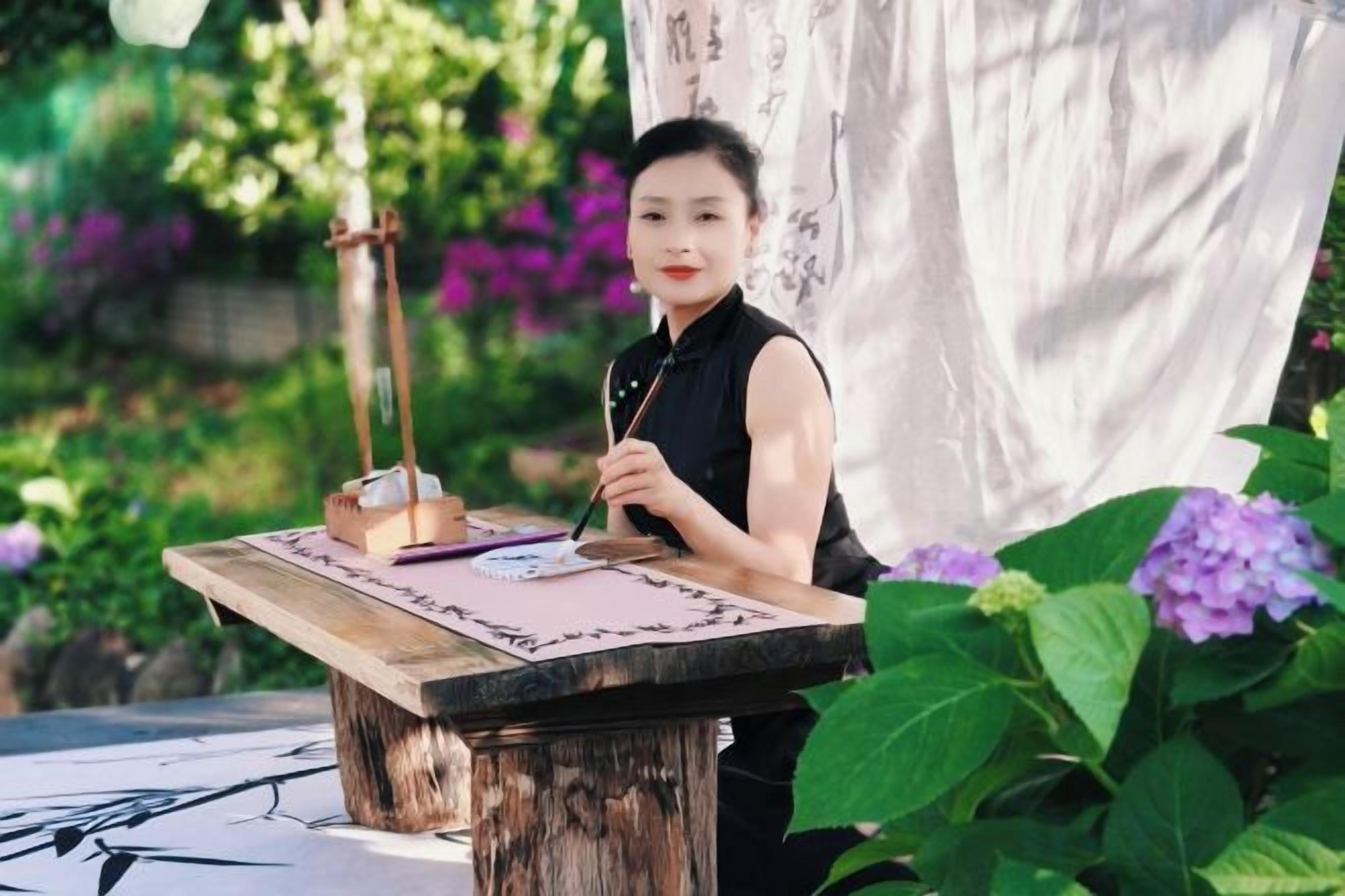 A person seated at a wooden table outdoors with a writing instrument and paper, surrounded by greenery and flowers.