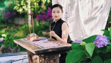 A person seated at a wooden table outdoors with a writing instrument and paper, surrounded by greenery and flowers.