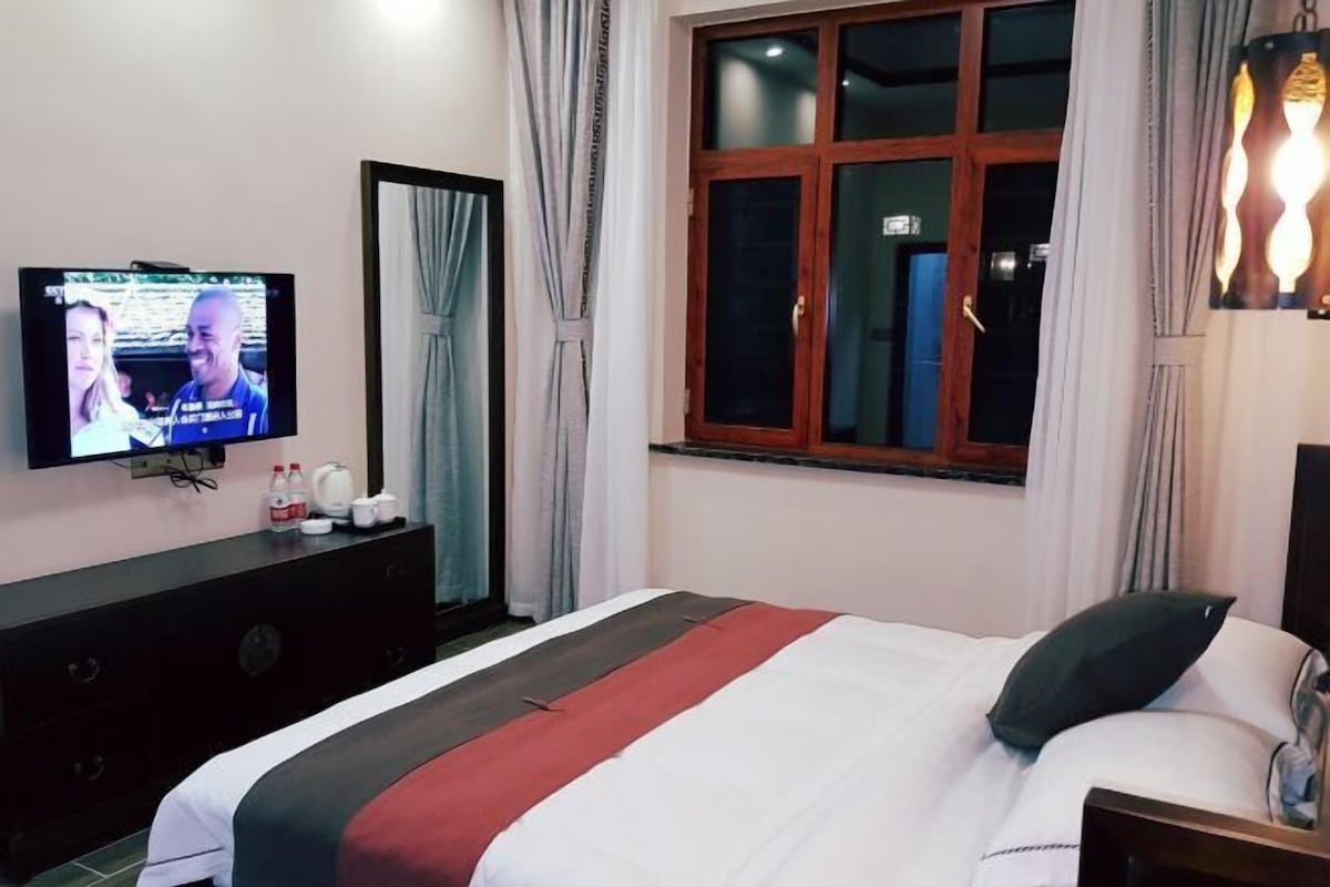 Una habitación de hotel con una cama, televisión en la pared, cómoda y ventana con cortinas.