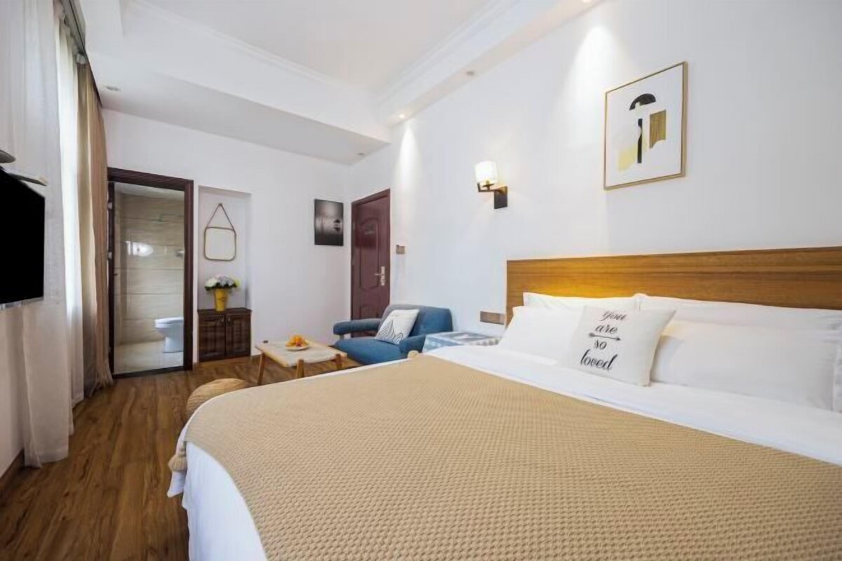 Habitación de hotel con una cama grande, suelo de madera, un sillón azul, una mesita con un florero y una puerta que da al baño.