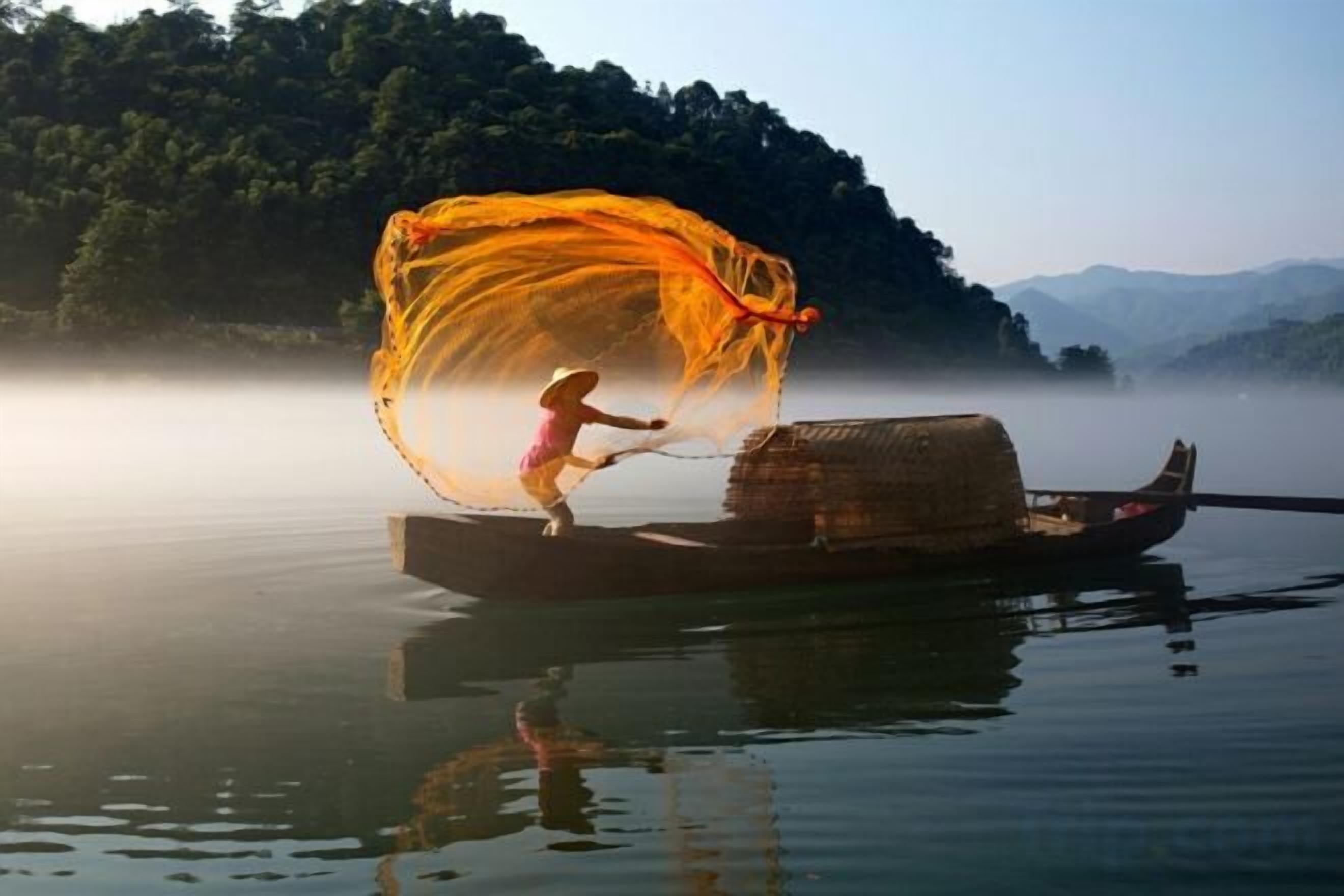 A person standing on a boat with a net, surrounded by calm water and a misty landscape.