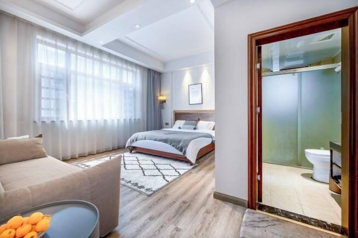 Ein modernes Hotelzimmer mit Bett, einer Couch, einem Esstisch mit Obst und einem durch eine Glastür sichtbaren Badezimmer.