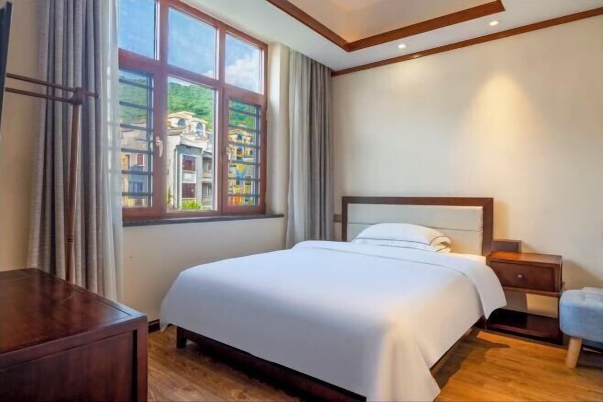 Una habitación de hotel con cama, mesita de noche, escritorio de madera, ventana con cortinas y vistas a edificios.