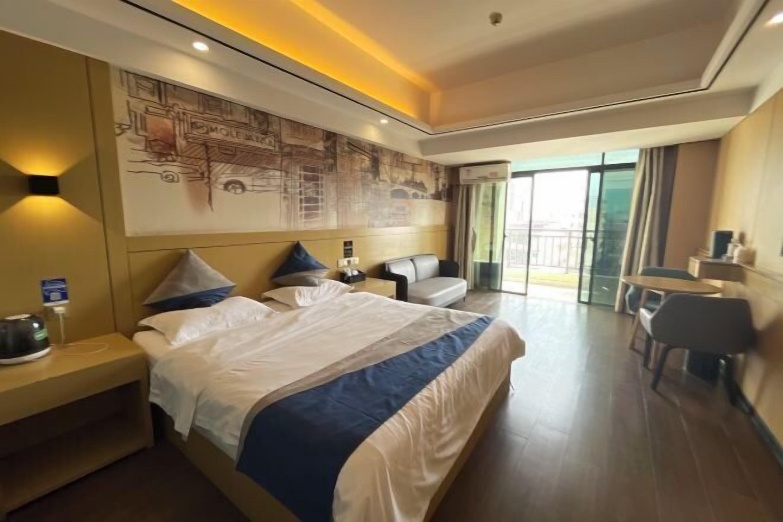A modern hotel room with a large bed, a seating area with a sofa and armchairs, a small table, and a large window offering a view of the city.