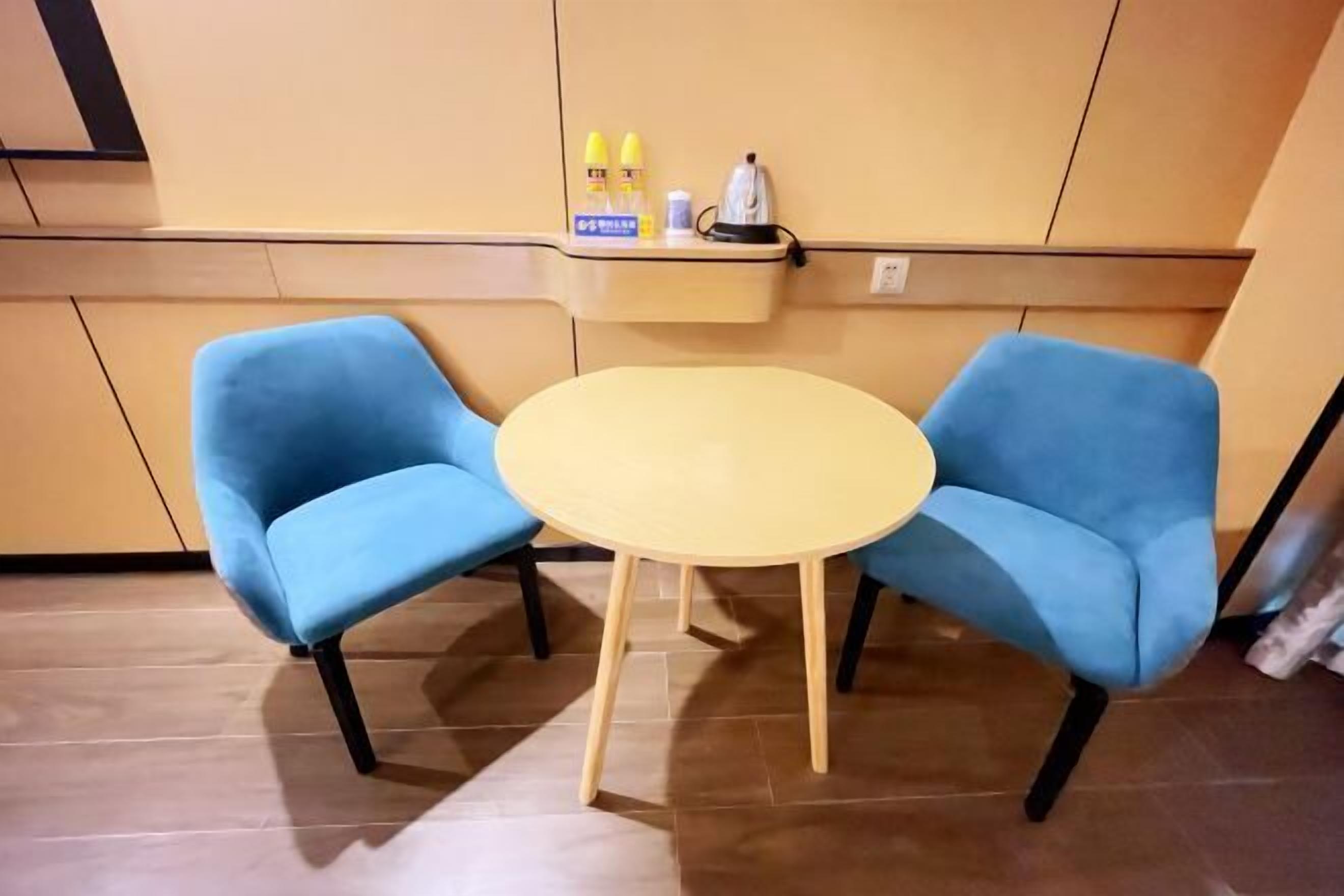 A room with two blue chairs around a small round table, a shelf with toiletries, and a wall-mounted kettle.